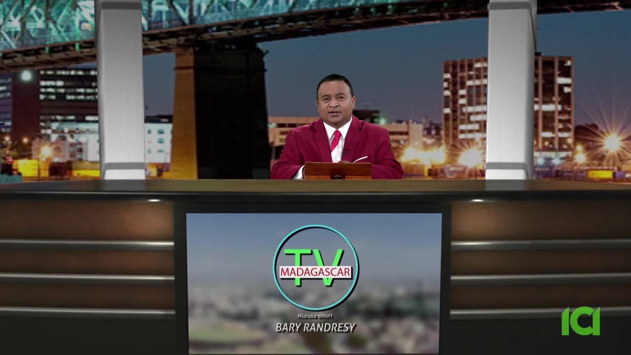 A man in a red suit sits behind a desk, speaking. Behind him, a cityscape glows under a dark sky, and a screen displays the "TV Madagascar" logo.
A man in a red suit sits behind a desk, speaking. Behind him, a cityscape glows under a dark sky, and a screen displays the "TV Madagascar" logo.