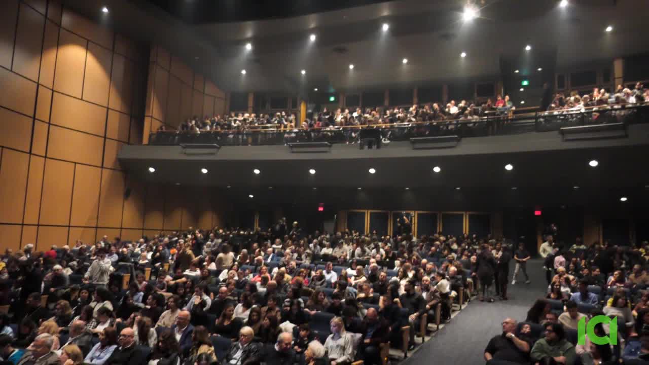 A large crowd fills the auditorium, with people seated in rows and more on the balcony above. The stage is empty, and the lighting is focused on the audience.
A large crowd fills the auditorium, with people seated in rows and more on the balcony above. The stage is empty, and the lighting is focused on the audience.