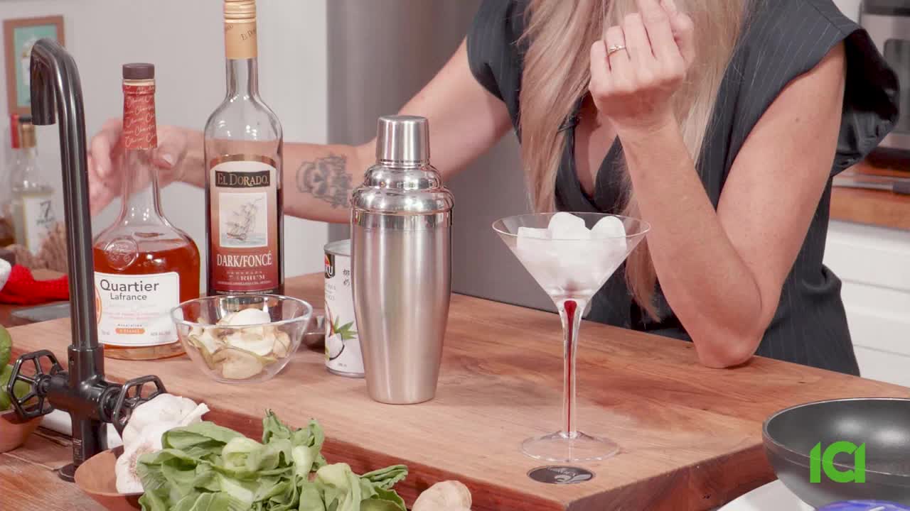 A woman is about to add something to a cocktail shaker, ingredients are laid out on the wooden counter. A martini glass filled with ice cubes sits ready.
