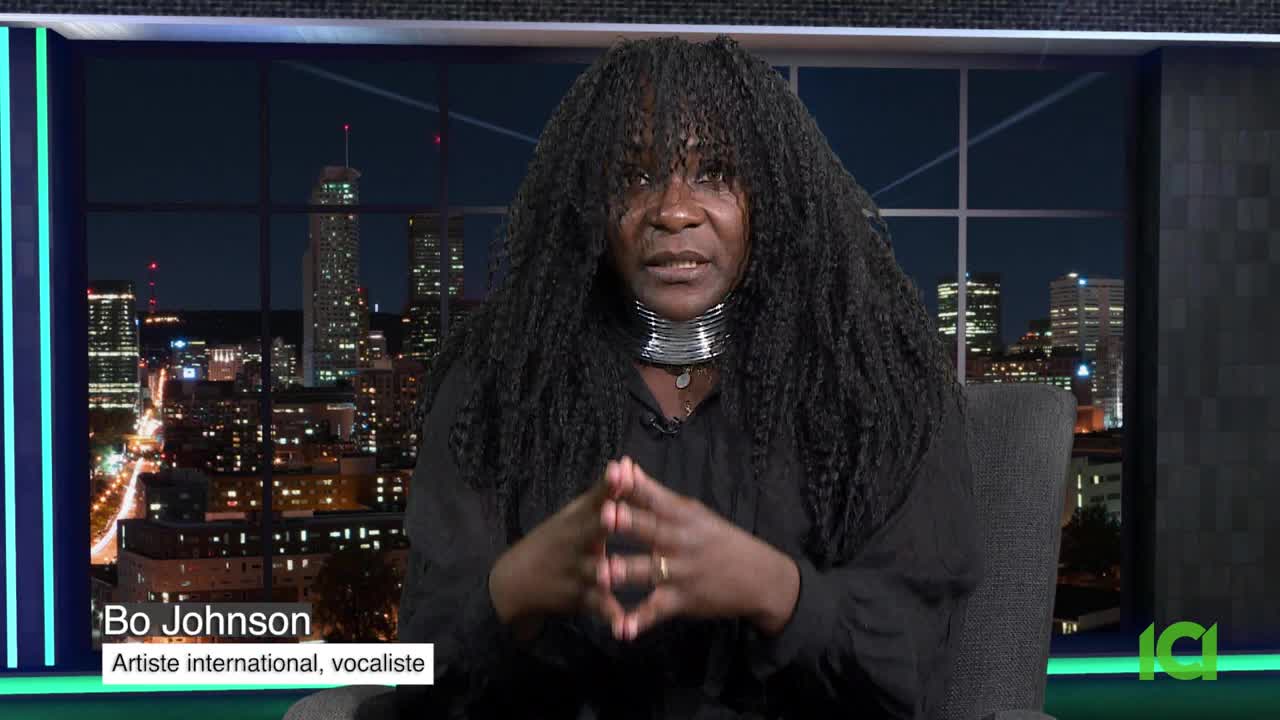 Bo Johnson, identified as an international artist and vocalist, sits in a studio chair, hands clasped, speaking on CFHD-DT in Canada. Behind her, a nighttime cityscape is visible through a large window.
