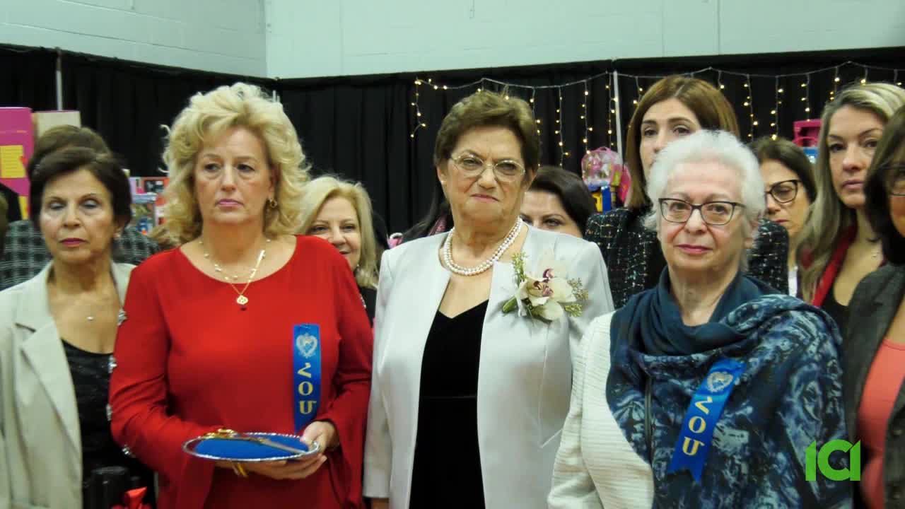A group of women in formal attire are gathered, some with blue ribbons pinned to their outfits. The woman in the center, wearing a white suit, looks directly at the camera while others stand behind her.
