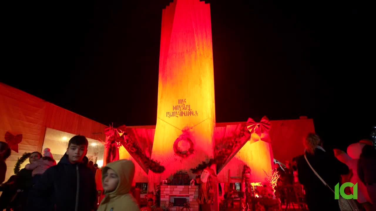 A tall, wooden structure glows with red and orange light, adorned with festive bows. People gather around, their faces illuminated by the vibrant display.