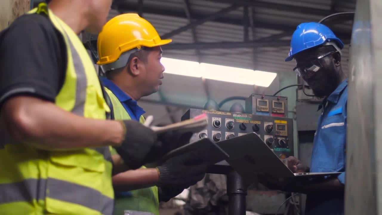 Three workers in hard hats and high-visibility vests gather around a control panel. One man in a blue uniform is focused on a laptop, while the others consult a tablet and a binder.