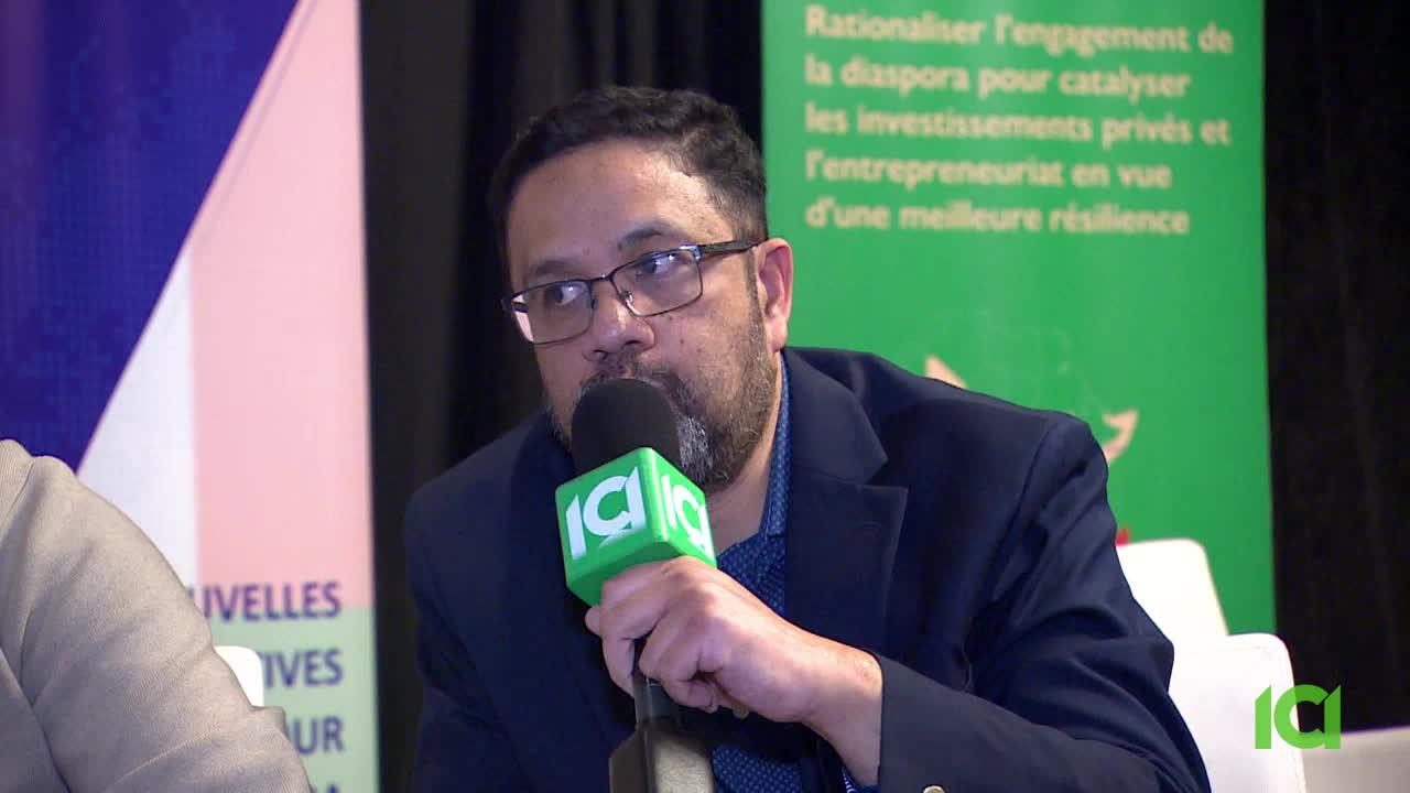 A man speaks into a bright green microphone, his hand resting on its stem. Behind him, a banner in French discusses diaspora engagement.