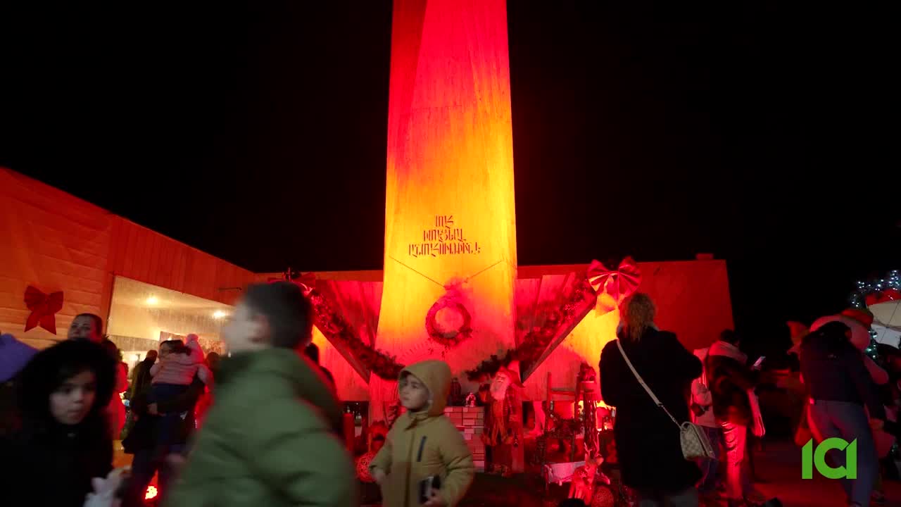 A tall, illuminated structure glows with warm red and yellow light, casting a festive atmosphere over a crowd gathered in the Canadian night. People mill about, some holding children, as they take in the holiday decorations.