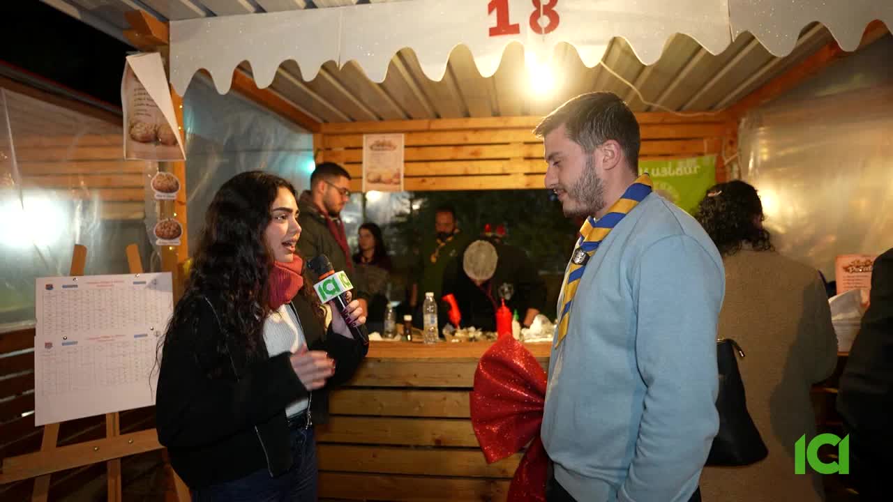 A young woman holds a microphone, speaking into it as she interviews a man wearing a blue sweater and a yellow and blue scarf. Behind them, people mill about a wooden stall decorated with a large red bow.