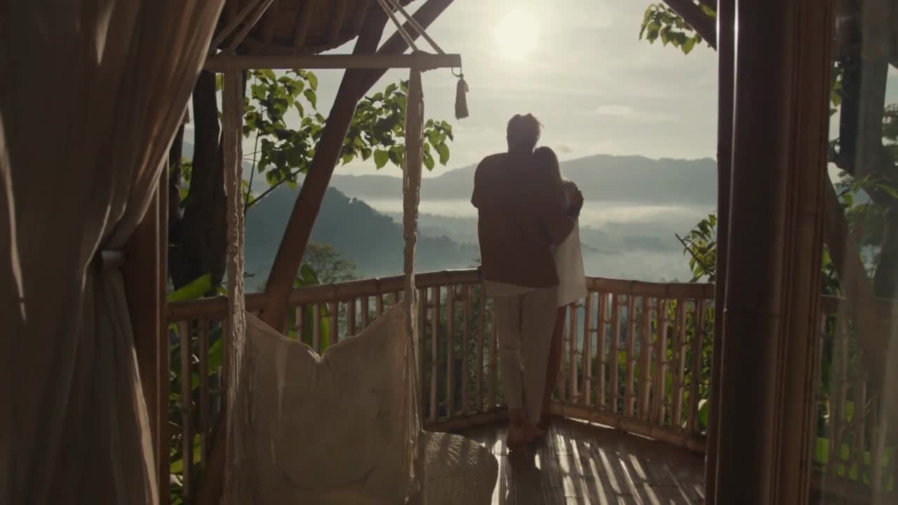 Two figures embrace on a wooden balcony, overlooking a misty valley bathed in morning light. Sunlight streams through the trees, casting long shadows across the deck.