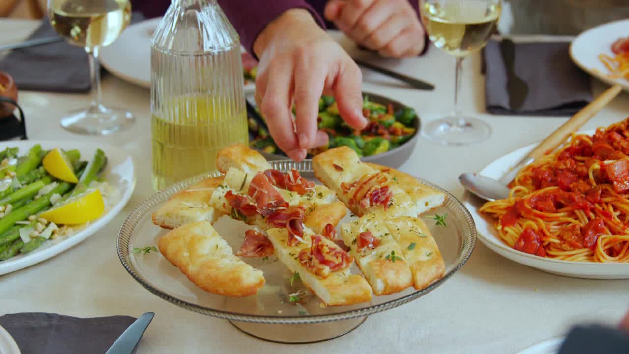 A hand reaches for a piece of flatbread topped with prosciutto. Beside it, a plate of spaghetti with a rich tomato sauce sits ready to be served.