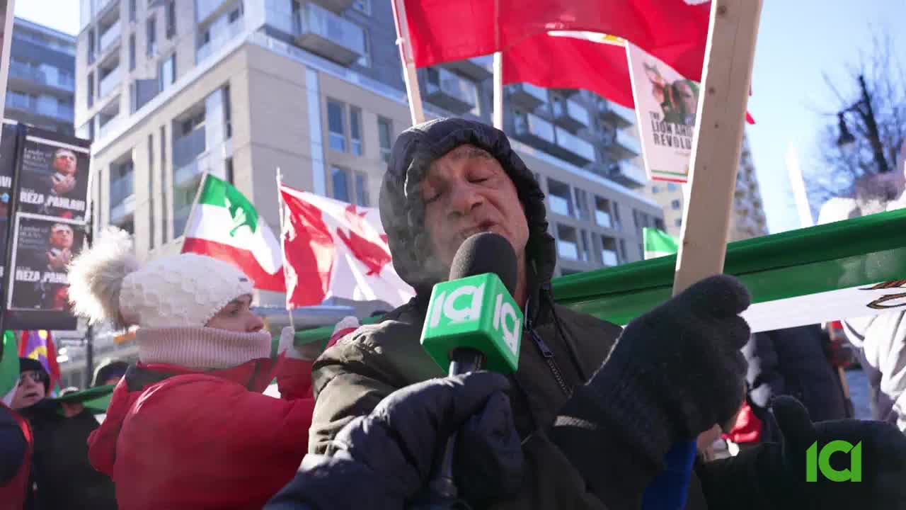 A man, speaking into a microphone with an ICI logo, stands amidst a crowd holding Iranian and Canadian flags. His breath mists in the cold air as he addresses the gathering.