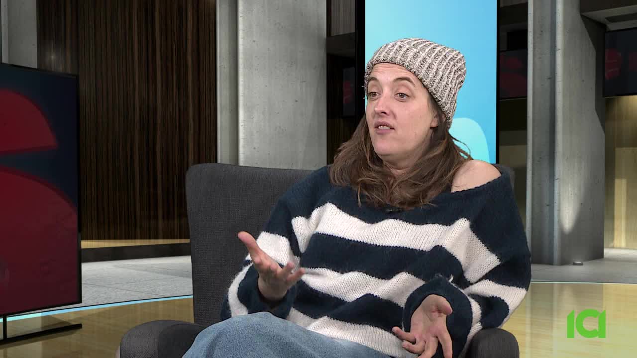 A woman in a striped sweater and beanie gestures with her hands while speaking. She's on a set that looks like it's for a broadcast, possibly for CFHD-DT in Canada.