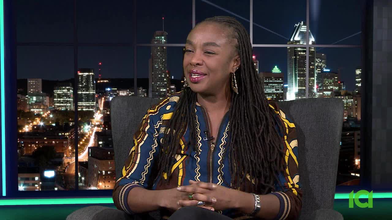 A woman with long, dark dreadlocks sits in a chair, her hands clasped in her lap. Behind her, the lights of Montreal twinkle against the night sky.