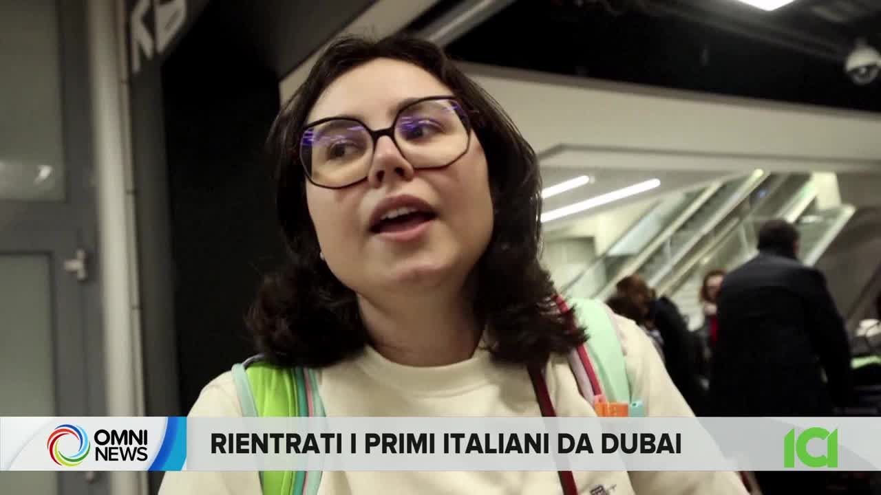 The young woman in glasses speaks into a microphone, her backpack slung over her shoulder. Behind her, people move past escalators in a busy terminal.