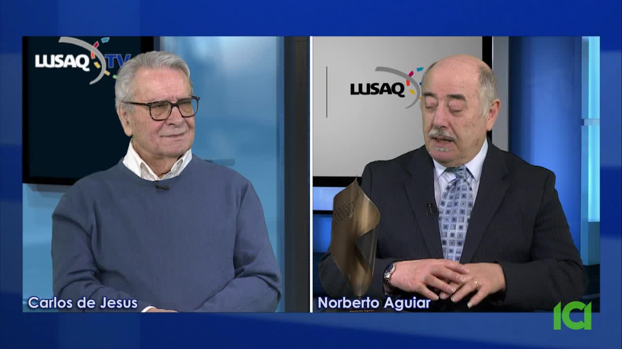 Two men are seated, speaking on a split screen. The man on the left, Carlos de Jesus, wears a blue sweater and glasses. Norberto Aguiar, on the right, is in a suit and tie, holding a bronze award.