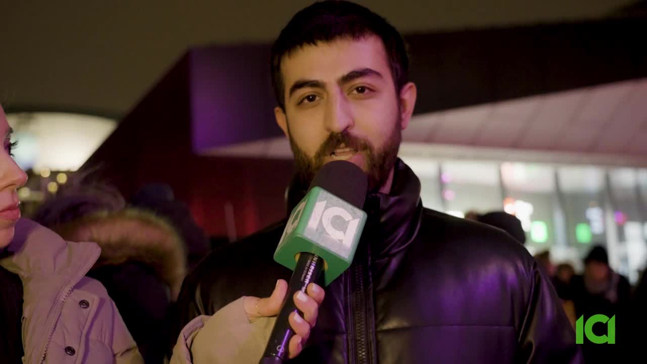A reporter holds an ICI microphone up to a man's face. The man is speaking into the mic, his beard visible under the bright lights.