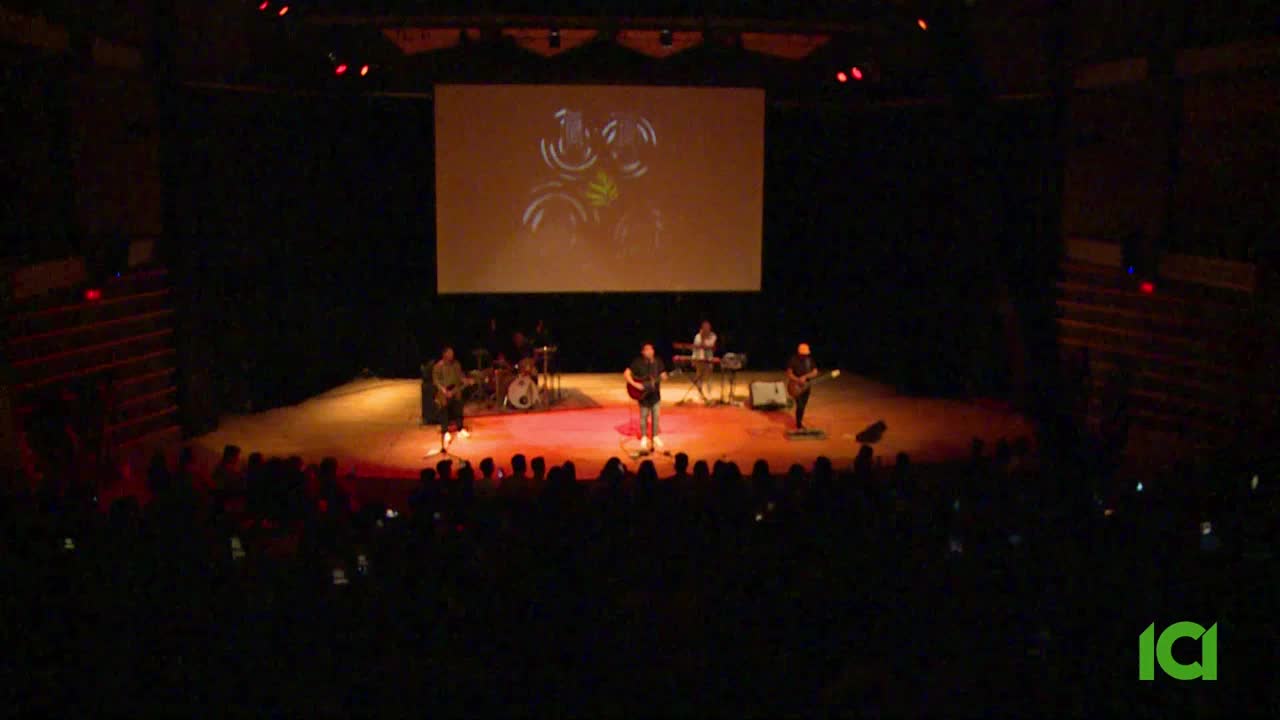 A band plays on stage in front of a large screen displaying abstract visuals. The audience, illuminated by their phones, watches the performance.