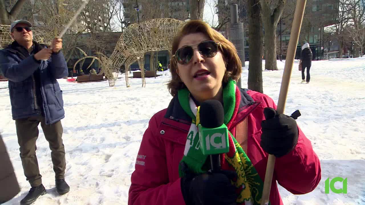 A reporter from CFHD-DT stands in the snow, holding a microphone and a wooden pole. Behind her, a man in a blue jacket holds a similar pole, and a large, wireframe deer decoration stands in the park.