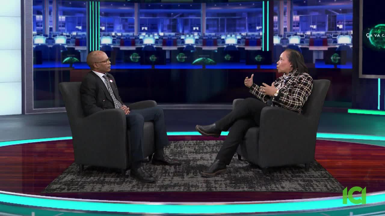 Two men sit in armchairs on a studio set, facing each other. The man on the left wears a dark suit and tie, while the man on the right has on a plaid jacket.