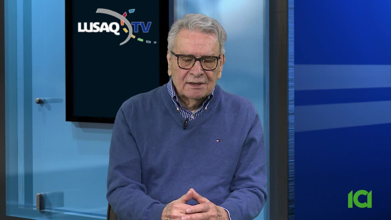 An older gentleman with glasses sits in front of a blue backdrop, a "LUSAQ TV" logo visible behind him. He wears a blue sweater over a collared shirt, his hands clasped in his lap. An older gentleman with glasses sits in front of a blue backdrop, a "LUSAQ TV" logo visible behind him. He wears a blue sweater over a collared shirt, his hands clasped in his lap.