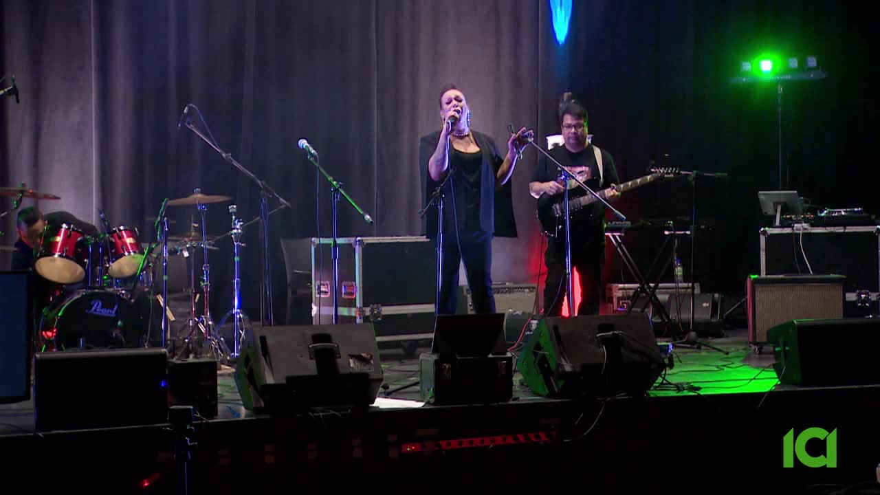 A woman sings into a microphone on a stage, her hand raised as a guitarist plays beside her. The band is set up with drums and keyboards behind them, bathed in colored stage lights. A woman sings into a microphone on a stage, her hand raised as a guitarist plays beside her. The band is set up with drums and keyboards behind them, bathed in colored stage lights.