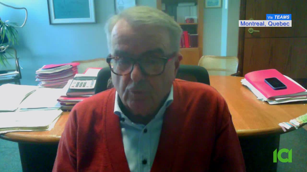 A man in a red cardigan speaks from his Montreal office. A stack of pink folders with a phone rests on his desk. A man in a red cardigan speaks from his Montreal office. A stack of pink folders with a phone rests on his desk.