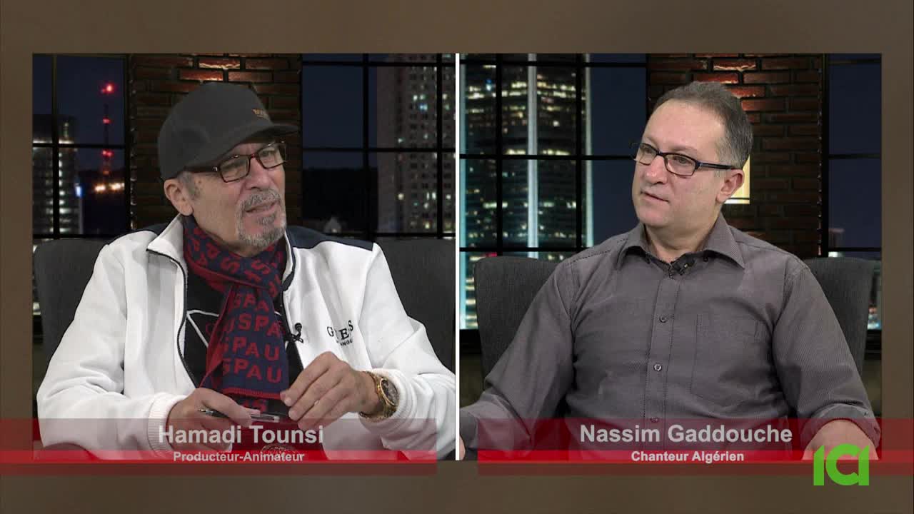 Two men sit in chairs, bathed in the warm glow of studio lights. The man on the left, wearing a cap and a patterned scarf, holds a pen. To his right, a man in a grey collared shirt looks thoughtfully ahead. Two men sit in chairs, bathed in the warm glow of studio lights. The man on the left, wearing a cap and a patterned scarf, holds a pen. To his right, a man in a grey collared shirt looks thoughtfully ahead.
