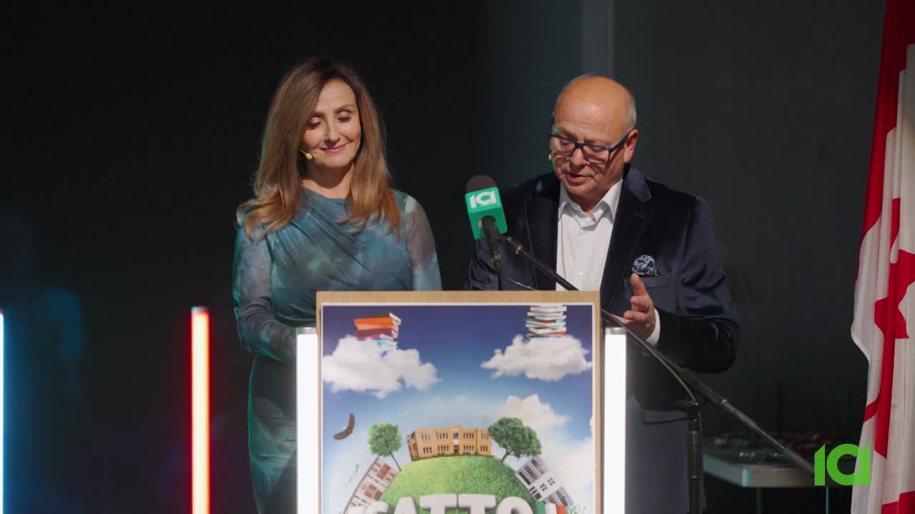 A man in a dark velvet jacket speaks into a microphone, gesturing with his hands. Beside him, a woman in a teal dress looks down, smiling gently. Behind them, a Canadian flag hangs, its red and white stark against the dark backdrop. A man in a dark velvet jacket speaks into a microphone, gesturing with his hands. Beside him, a woman in a teal dress looks down, smiling gently. Behind them, a Canadian flag hangs, its red and white stark against the dark backdrop.