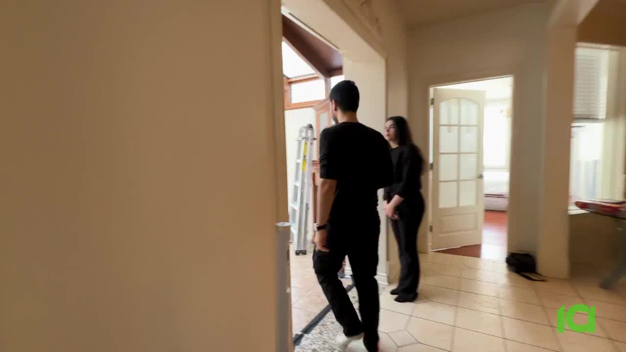 A man and woman stand in a hallway, looking towards a sunlit room. A silver ladder leans against the wall near them. A man and woman stand in a hallway, looking towards a sunlit room. A silver ladder leans against the wall near them.