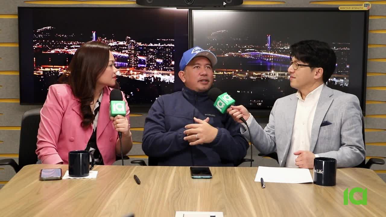 A woman in a pink blazer and two men, one in a blue jacket and baseball cap, the other in a grey suit, sit at a table holding microphones. They are in a studio with a city skyline displayed on screens behind them, likely for a broadcast from CFHD-DT in Canada.