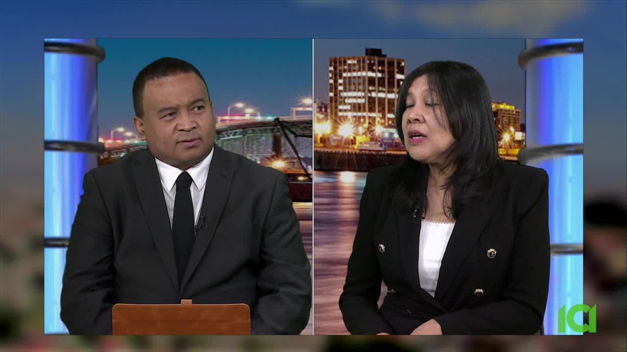 Two people, a man and a woman, are seated in front of a backdrop of a city skyline at night. The man, wearing a suit and tie, looks off to the side. The woman, in a black blazer, speaks with her eyes closed.