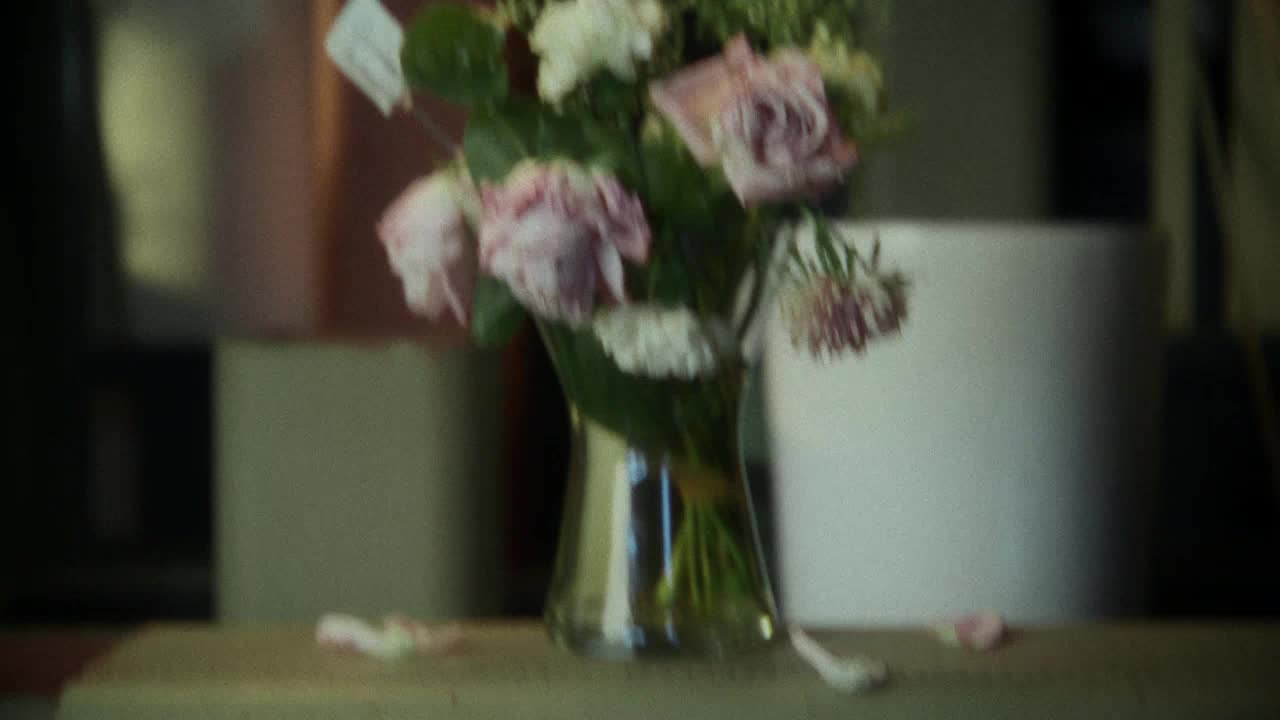 A bouquet of wilting pink and white roses sits in a glass vase. A few petals have fallen onto the surface below.