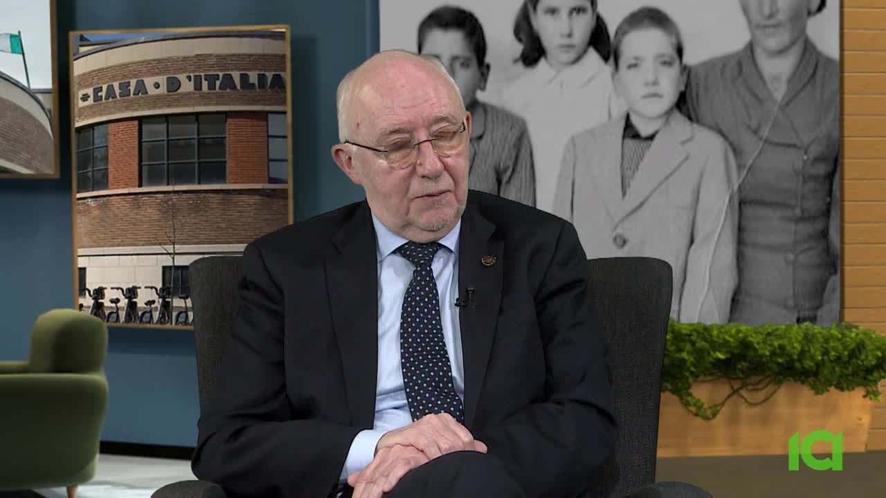 An older man in a suit sits in a chair, speaking. Behind him, a black and white photograph shows a group of children. To the left, a framed picture displays a brick building with "CASA D'ITALIA" written above its windows.