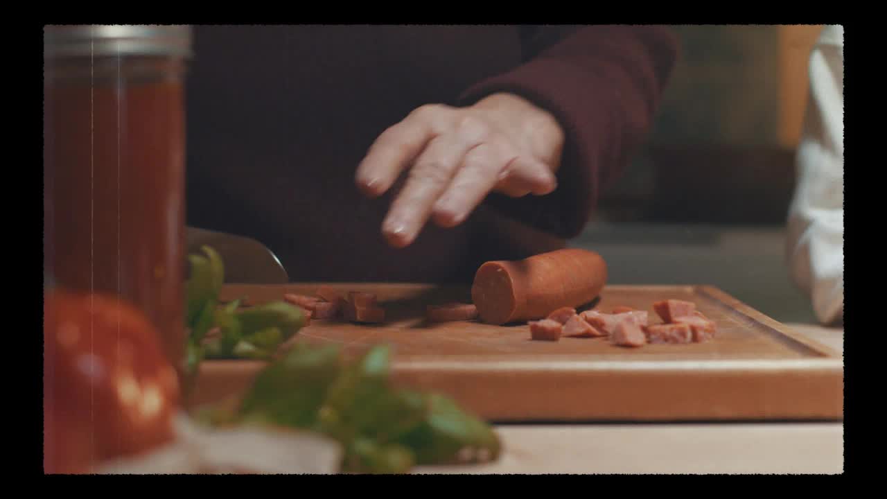 A hand moves over a cutting board, slicing a sausage into small cubes. A jar of tomato sauce sits to the left, and fresh herbs are scattered nearby.