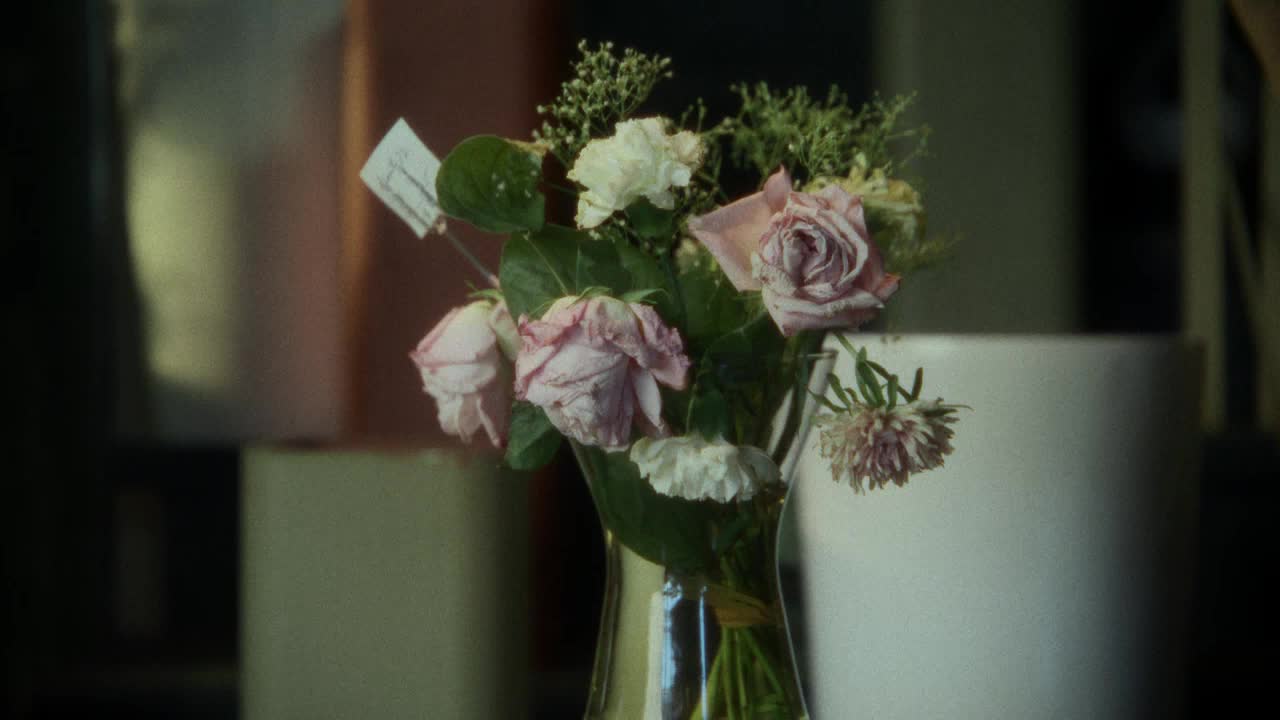 A bouquet of wilting roses sits in a glass vase. A small white tag is attached to one of the stems.