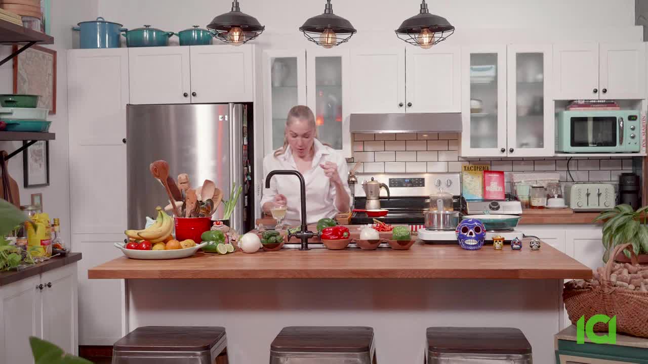 A woman in a white shirt is pouring liquid from a small bottle into a glass at a kitchen island. Colorful produce and a blue sugar skull sit on the counter before her.