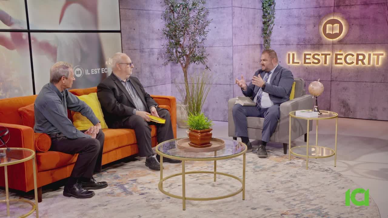 The man in the suit is gesturing with his hands as he speaks. Beside him, another man sits on an orange couch, holding a yellow book.