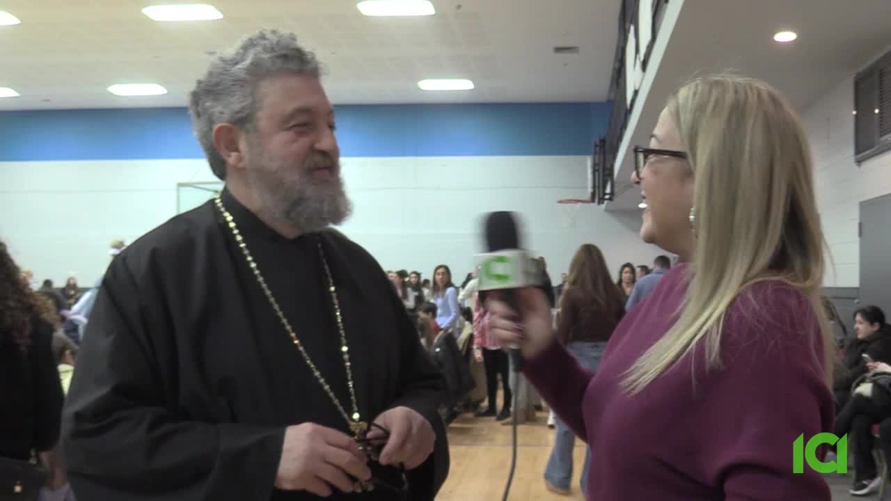 A reporter from CFHD-DT interviews a man in traditional Orthodox attire in a gymnasium. People mill about in the background of this Canadian event.
