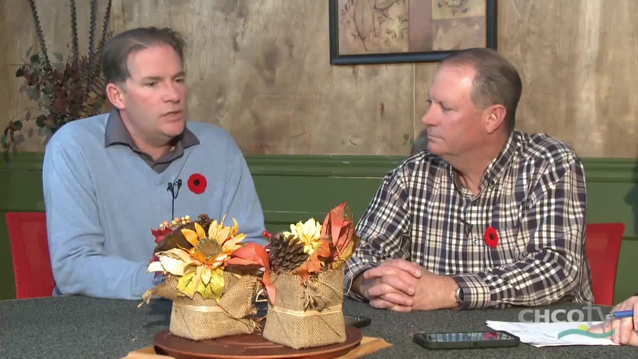 Two men, each wearing a poppy, are seated at a table, likely on a CHCO-TV set in Canada. The man on the left is speaking, while the other listens intently.
