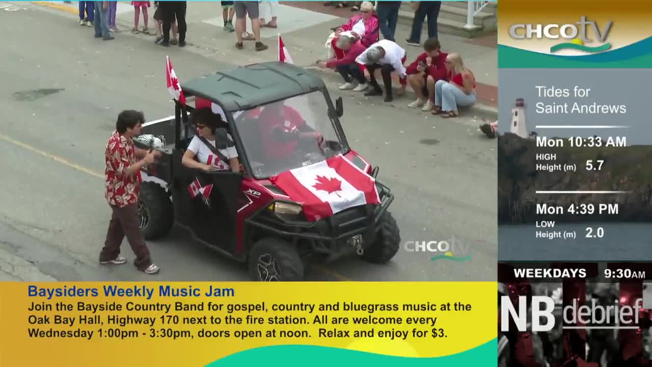 A small ATV, draped with a Canadian flag, moves down the street. A man in a Hawaiian shirt gestures toward the vehicle, as people watch from the side.
