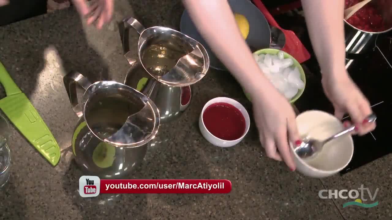 A person is stirring something white in a small bowl, with a spoon. Beside them, there are bowls of ice, red liquid, and metal pitchers filled with a clear liquid.
