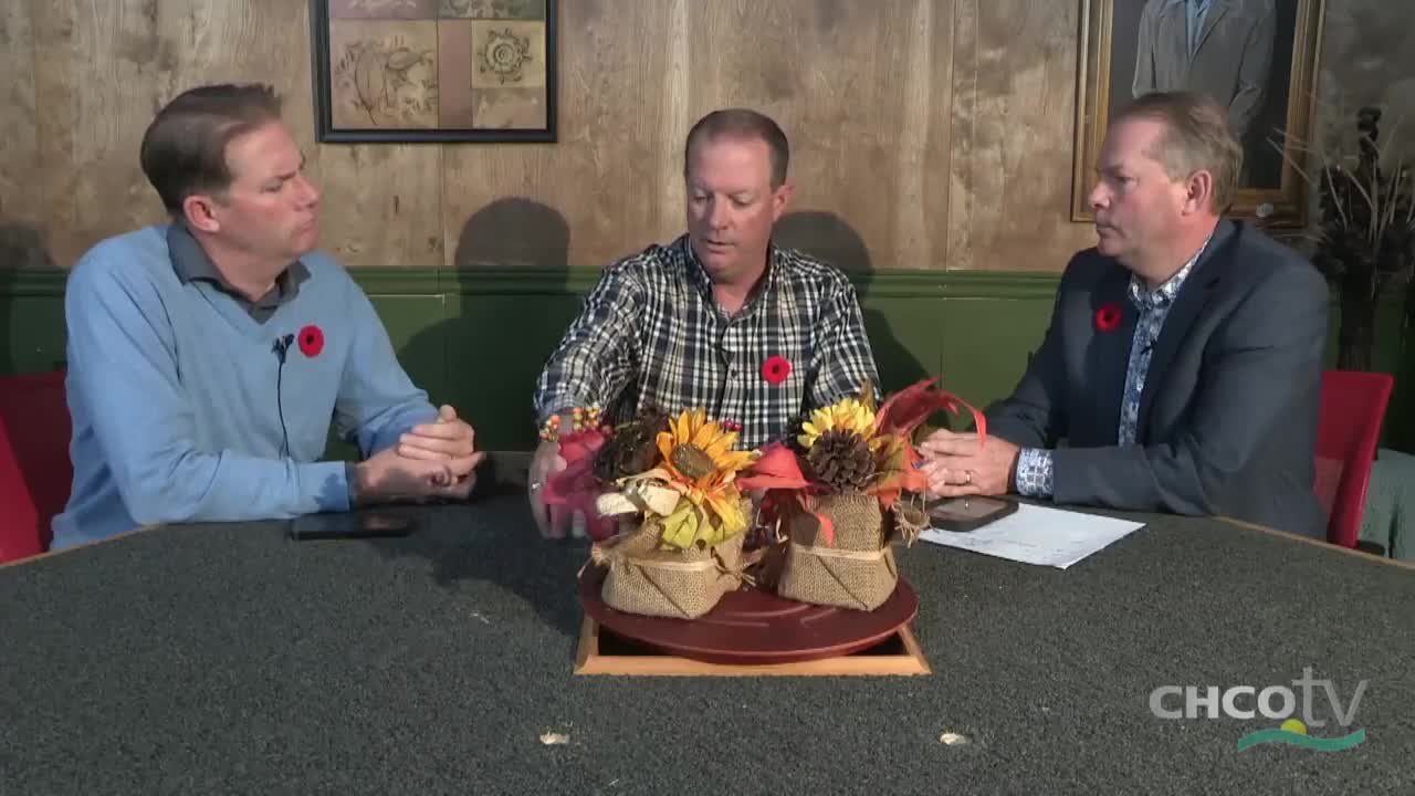 Three men sit around a table, each wearing a poppy. The man in the center gestures toward a small floral arrangement.
