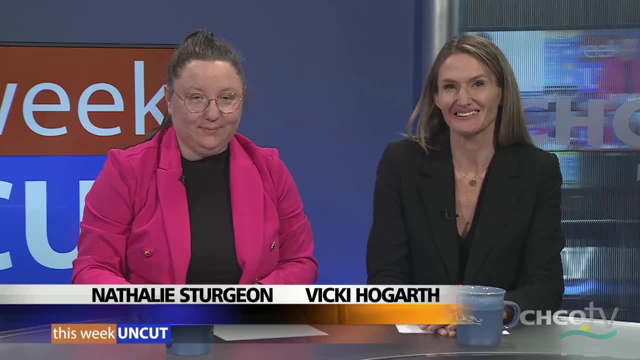 Two women sit at a desk, smiling at the camera on the CHCO-TV set. The woman on the left wears a bright pink blazer, while the other is in a black jacket.
