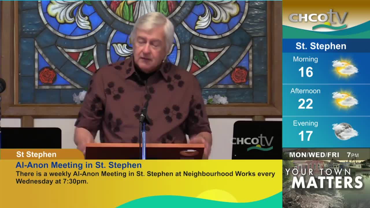 A man with silver hair stands at a podium, speaking into a microphone. Beside him, the CHCO-TV weather forecast for St. Stephen, Canada, is displayed.
