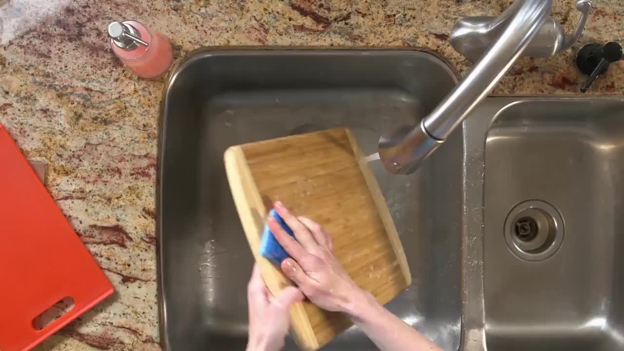 A wooden cutting board is being scrubbed with a blue sponge under running water in a stainless steel sink. Beside it, a red cutting board sits on the counter.

