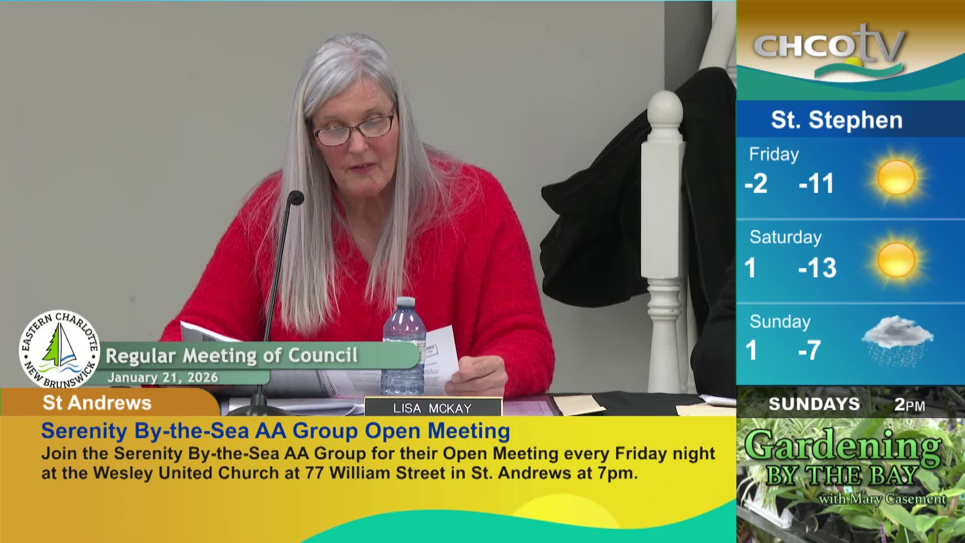 A woman with long grey hair, wearing a bright red sweater, speaks at a table. A CHCO-TV weather report shows cold temperatures for St. Stephen, New Brunswick.