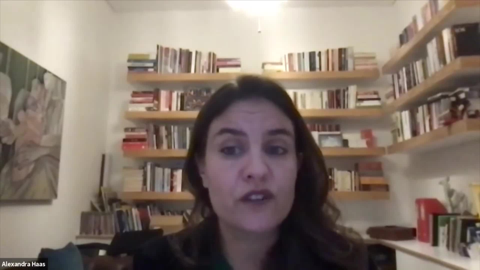 A woman is speaking directly to the camera, her expression earnest. Behind her, shelves overflow with books, a testament to a life of learning.