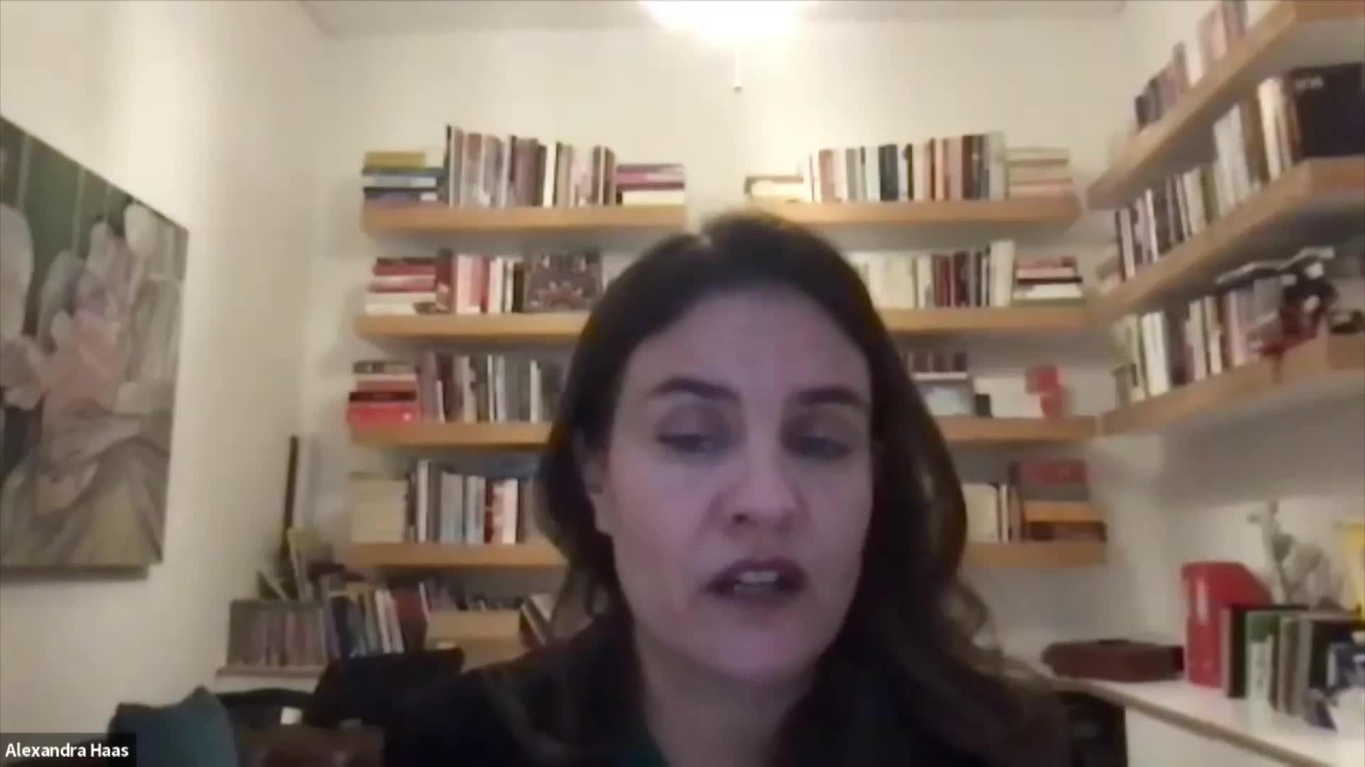 Alexandra Haas is speaking directly to the camera. Behind her, shelves packed with books line the wall.