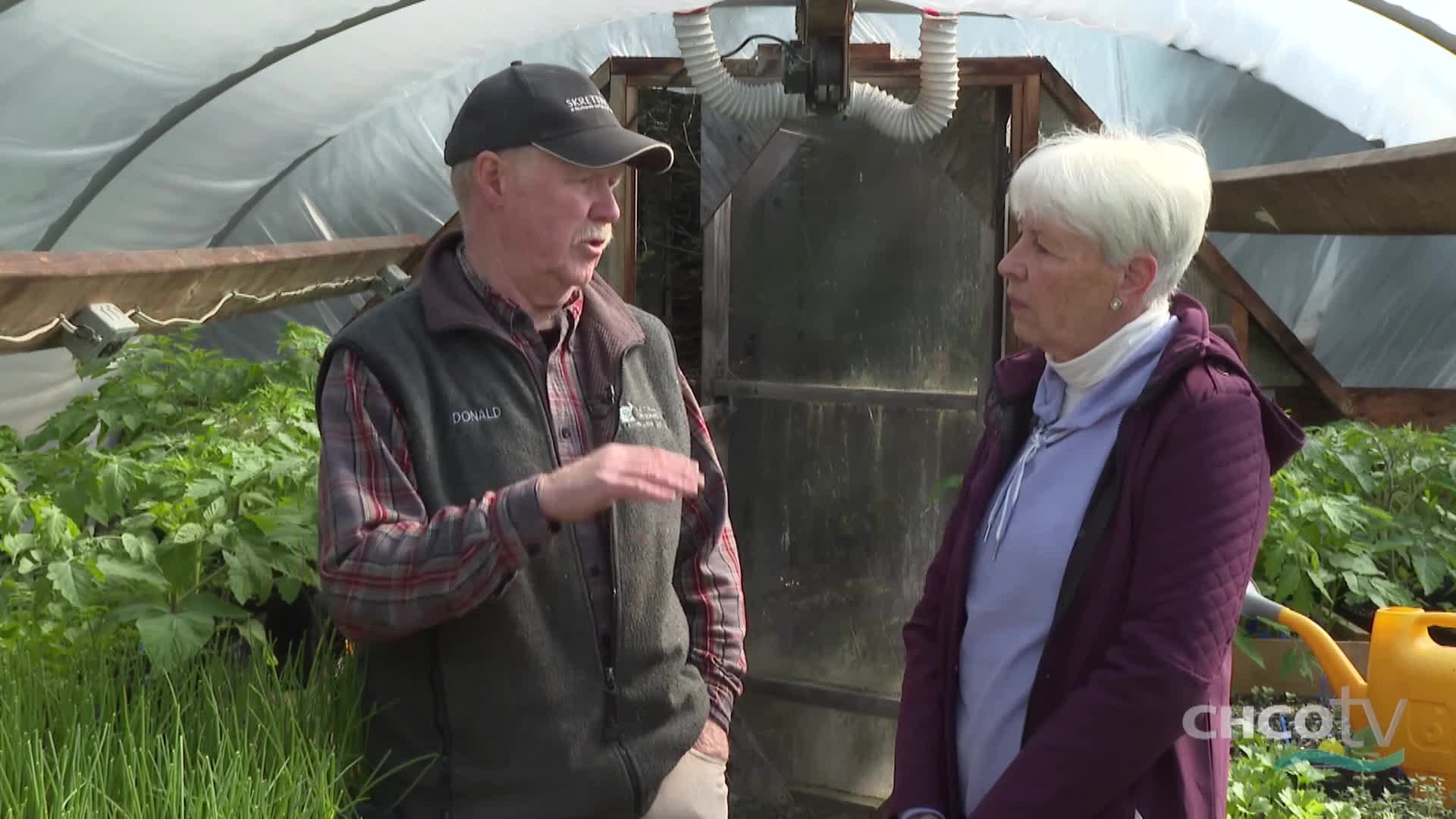 A man in a cap gestures with his hand while speaking to a woman inside a greenhouse. Lush green plants fill the space around them, with a yellow watering can visible in the background.