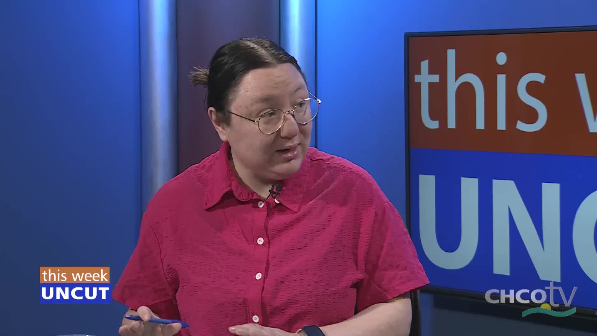A woman in a bright pink shirt gestures with a pen. Behind her, a screen displays "this week UNCUT" and the CHCO-TV logo.