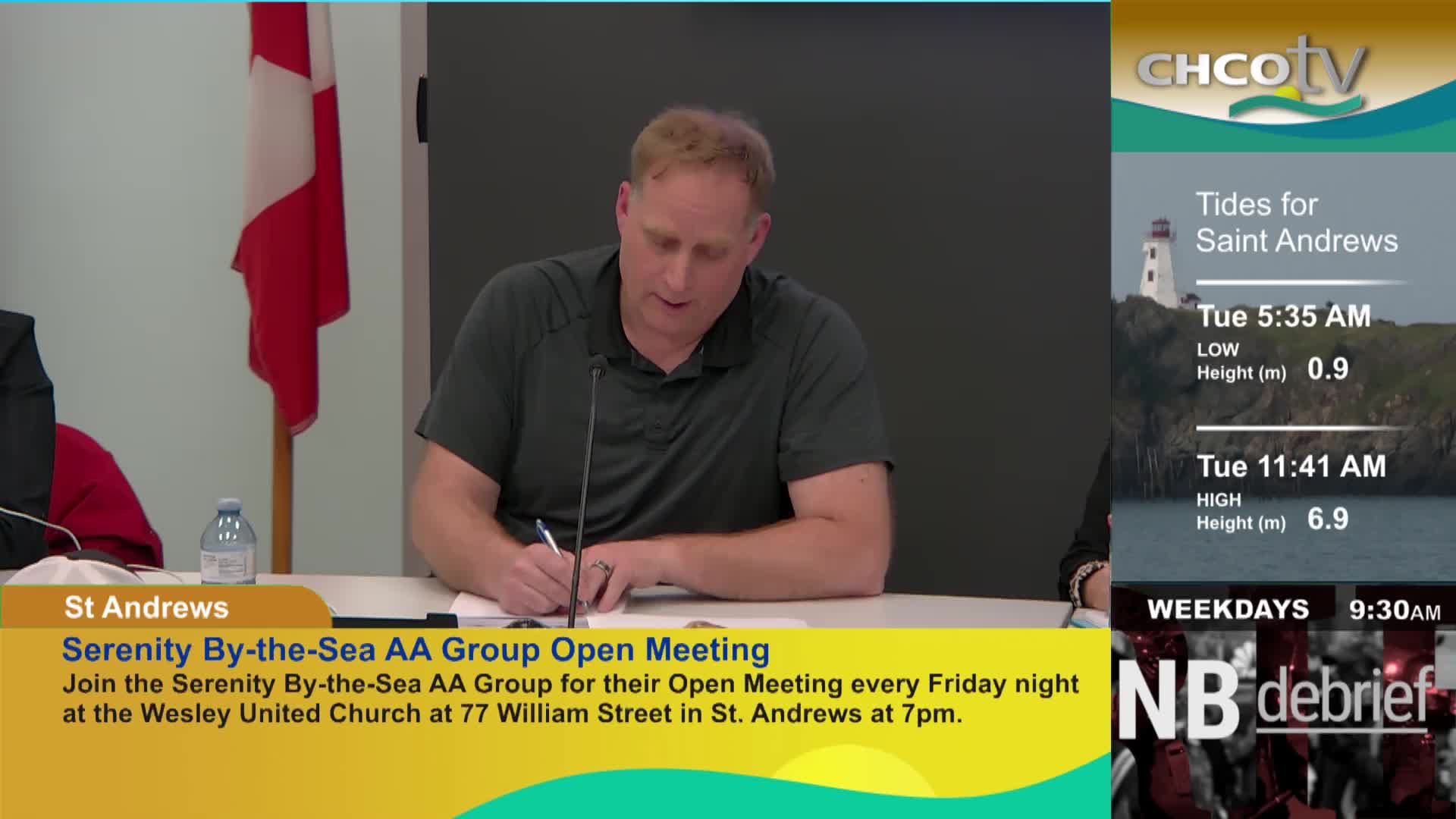 A man in a dark grey polo shirt leans over a table, pen in hand, writing on a document. Beside him, a Canadian flag hangs on the wall.
