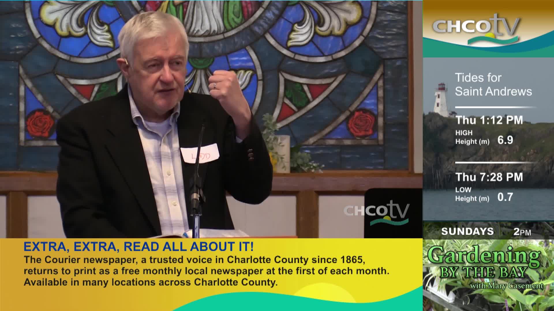 A man in a suit gestures with his right hand while speaking into a microphone. On the right side of the screen, CHCO-TV displays tide information for Saint Andrews and a gardening program. A man in a suit gestures with his right hand while speaking into a microphone. On the right side of the screen, CHCO-TV displays tide information for Saint Andrews and a gardening program.